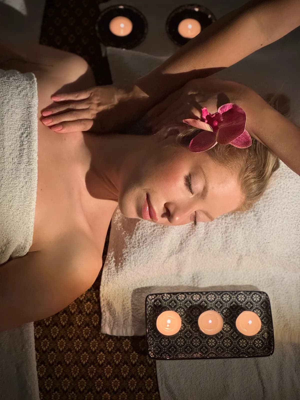 Sproščena stranka z zaprtimi očmi in orhidejo v laseh, medtem ko terapevtka med aromatično masažo ob svečah pritiska na njeno ramo